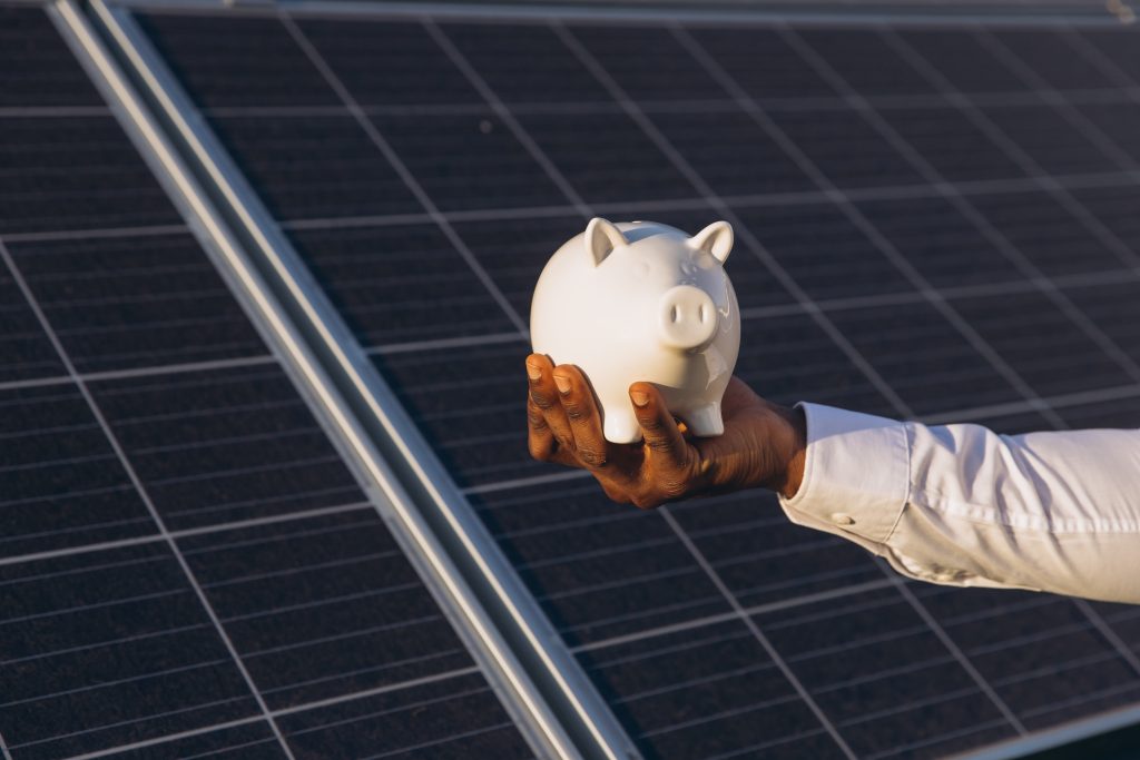 Hand holding a piggy bank next to solar panels, symbolising energy cost savings.